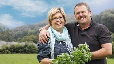 Lächelnder Mann und Frau stehen in einem grünen Feld vor bewaldeten Hügeln, halten frischen Basilikum aus Eigenproduktion und umarmen sich.