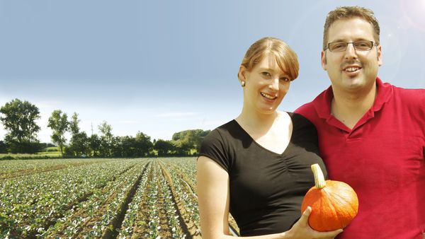 Lächelnder Mann und Frau stehen in einem sonnigen Kürbisfeld, halten einen orangefarbenen Kürbis aus Eigenproduktion. Im Hintergrund sind Reihen von Kürbispflanzen und Bäume zu sehen.