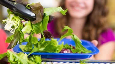 Eine Person gibt frischen Salat vom Foodservice mit einer Zange auf einen blauen Teller, im Hintergrund lächelt eine Frau