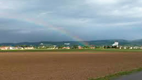 Weites braunes Feld vor einer Dorfkulisse und Hügeln, über denen ein Regenbogen am bewölkten Himmel erscheint.